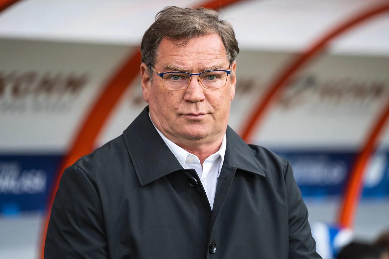 Jan Urban nowym selekcjonerem reprezentacji Polski Mężczyzna w okularach i ciemnym płaszczu stoi na tle trybun stadionu, patrząc prosto w obiektyw z poważnym wyrazem twarzy.