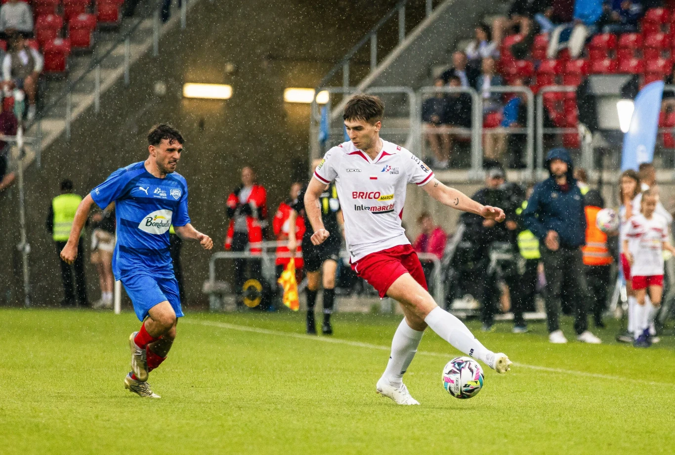 Dwóch piłkarzy rywalizuje o piłkę podczas meczu na stadionie, zawodnik w białym stroju wykonuje dalekie podanie, a w tle widoczni są kibice na trybunach oraz inni gracze.