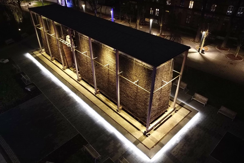 Drewniana tężnia solankowa oświetlona nocą, otoczona nowoczesnym chodnikiem i ławkami, widziana z góry, z jasnym pasem światła podkreślającym jej kształt.