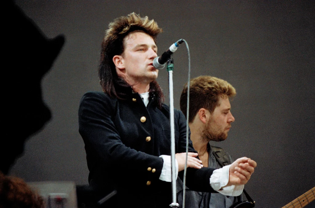 Bono podczas koncertu Live Aid Dwóch muzyków na scenie, jeden z nich śpiewa do mikrofonu, drugi gra na gitarze. Ubrani w ciemne ubrania, skupieni na występie, w tle prosty, neutralny sceniczny wystrój.