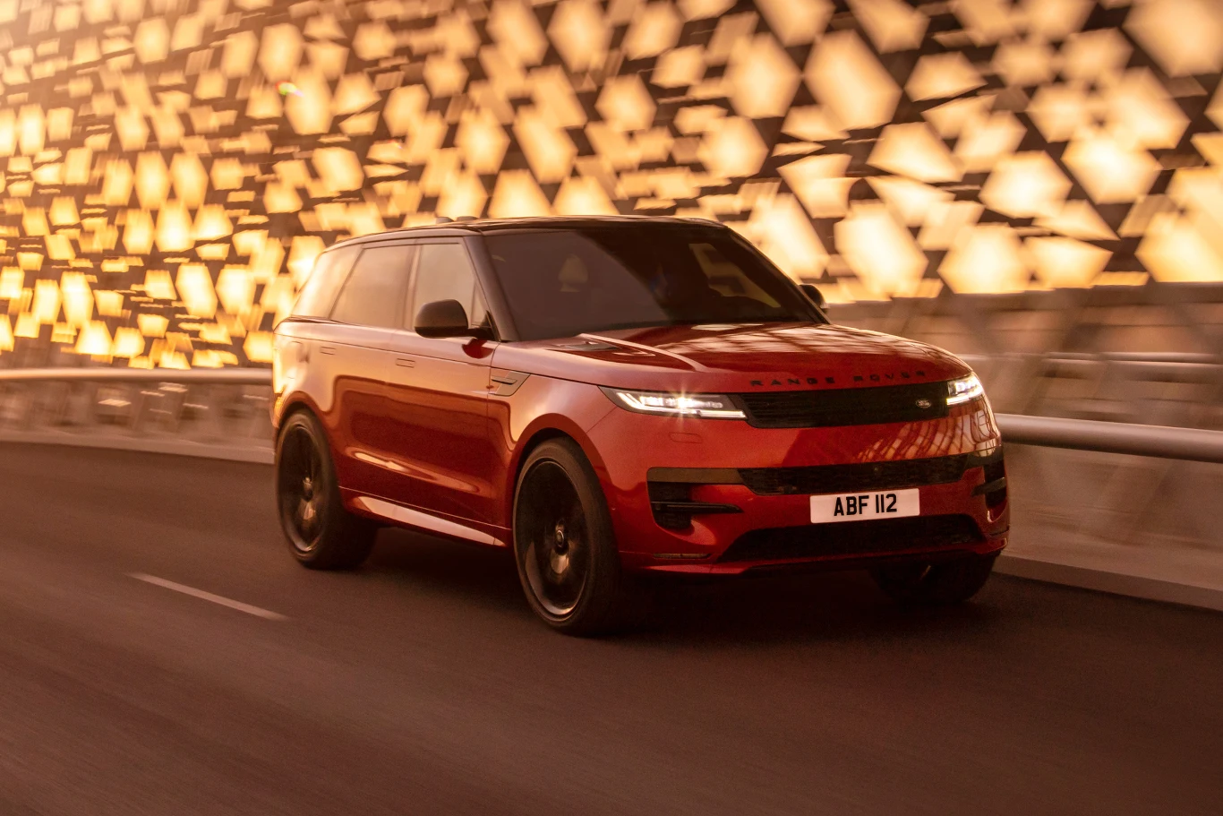 czerwony SUV marki Range Rover jadący dynamicznie po nowoczesnym moście, w tle abstrakcyjne, podświetlone wzory na ścianie