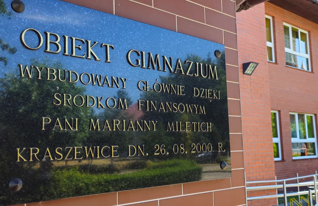Tablica informacyjna zamontowana na ścianie budynku szkoły, wykonana z czarnego kamienia z wyrytymi złotymi literami informującymi o powstaniu obiektu gimnazjum dzięki środkom finansowym konkretnej osoby, w tle fragment budynku z oknami.