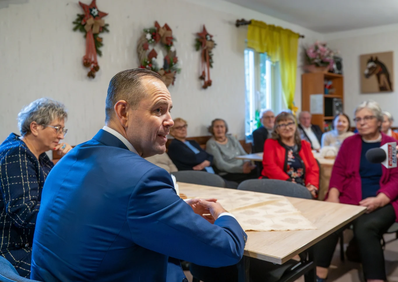 W czasie kampanii Karol Nawrocki obiecał, że w pierwszym dniu objęcia urzędu prezydenta złoży do Sejmu projekt ustawy zmieniające emerytury Grupa seniorów siedzi przy stołach w jasnym, udekorowanym kwiatami i wieńcami pomieszczeniu, jeden mężczyzna w garniturze zajmuje miejsce najbliżej kamery i rozmawia z uczestnikami spotkania