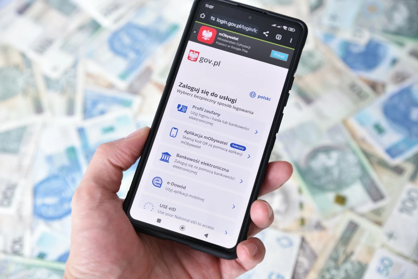 Nowa opcja w mObywatelu. Tusk o wielkiej zmianie. "Dla ludzi to bardzo poważny krok" Wyświetlony ekran logowania do platformy gov.pl na smartfonie trzymanym w dłoni, w tle rozłożone polskie banknoty o różnych nominałach.