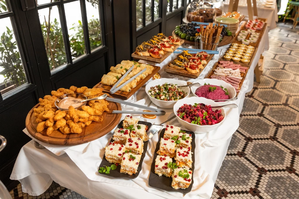 Bogato zastawiony bufet z różnorodnymi zakąskami, sałatkami, tartami, koreczkami i ciastami na białym obrusie, ustawiony przy dużych oknach w eleganckim wnętrzu restauracji.
