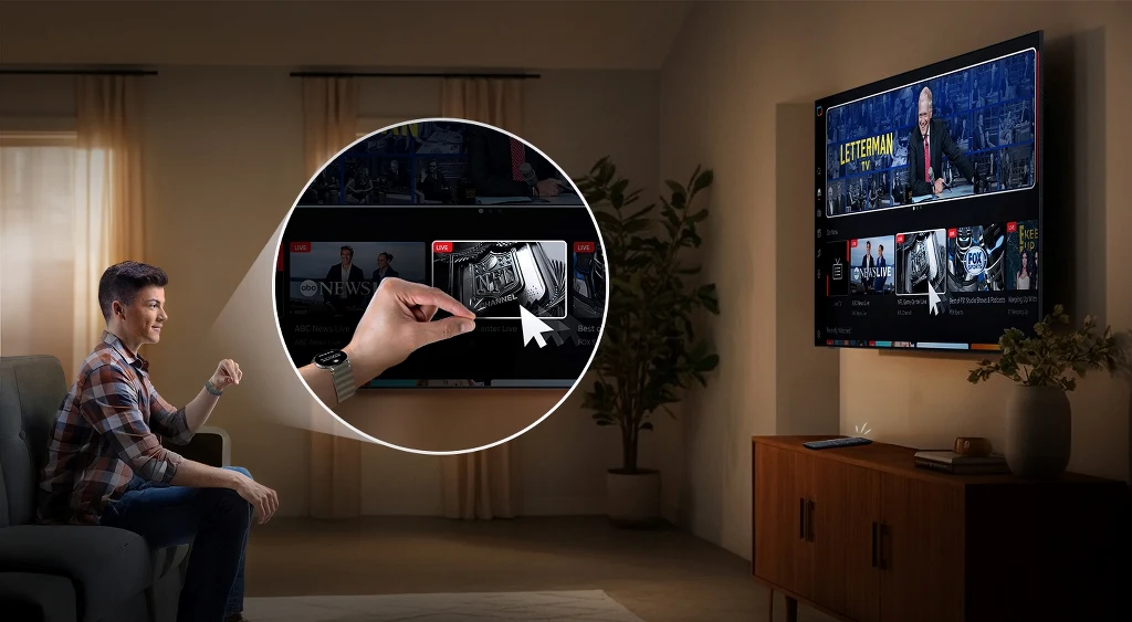 Tegoroczną linię telewizorów OLED spaja technologia Samsung Vision AI Osoba siedząca na sofie w salonie korzysta z telewizora z funkcją przeglądania i wybierania treści przy pomocy pilota. W pomieszczeniu znajduje się zabudowa z drewnianą komodą, telewizor zawieszony jest na ścianie, a duże okna są zasłonięte firanami. P...