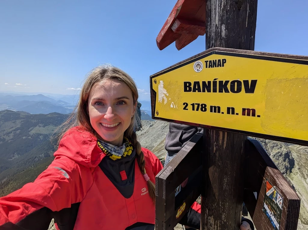 Baníkov, najwyższy szczyt Grani Rohaczy Uśmiechnięta kobieta w czerwonej kurtce robi selfie na szczycie górskim Baníkov, obok żółtego drogowskazu z nazwą i wysokością 2178 m n.p.m., w tle rozciągają się góry i błękitne niebo.