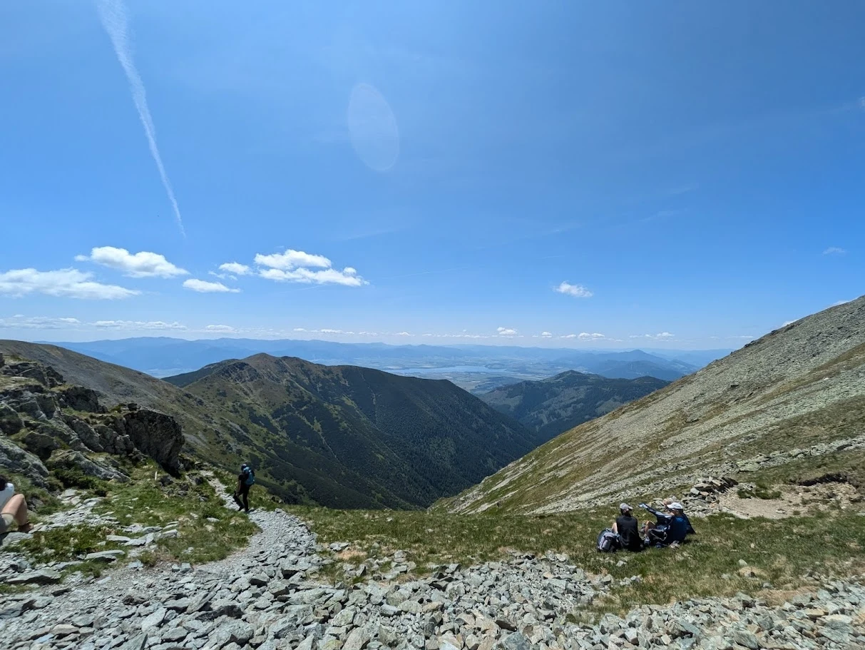 Szeroka panorama górska z zielonymi dolinami, niebieskim niebem oraz pojedynczymi osobami odpoczywającymi i idącymi po skalistym szlaku.