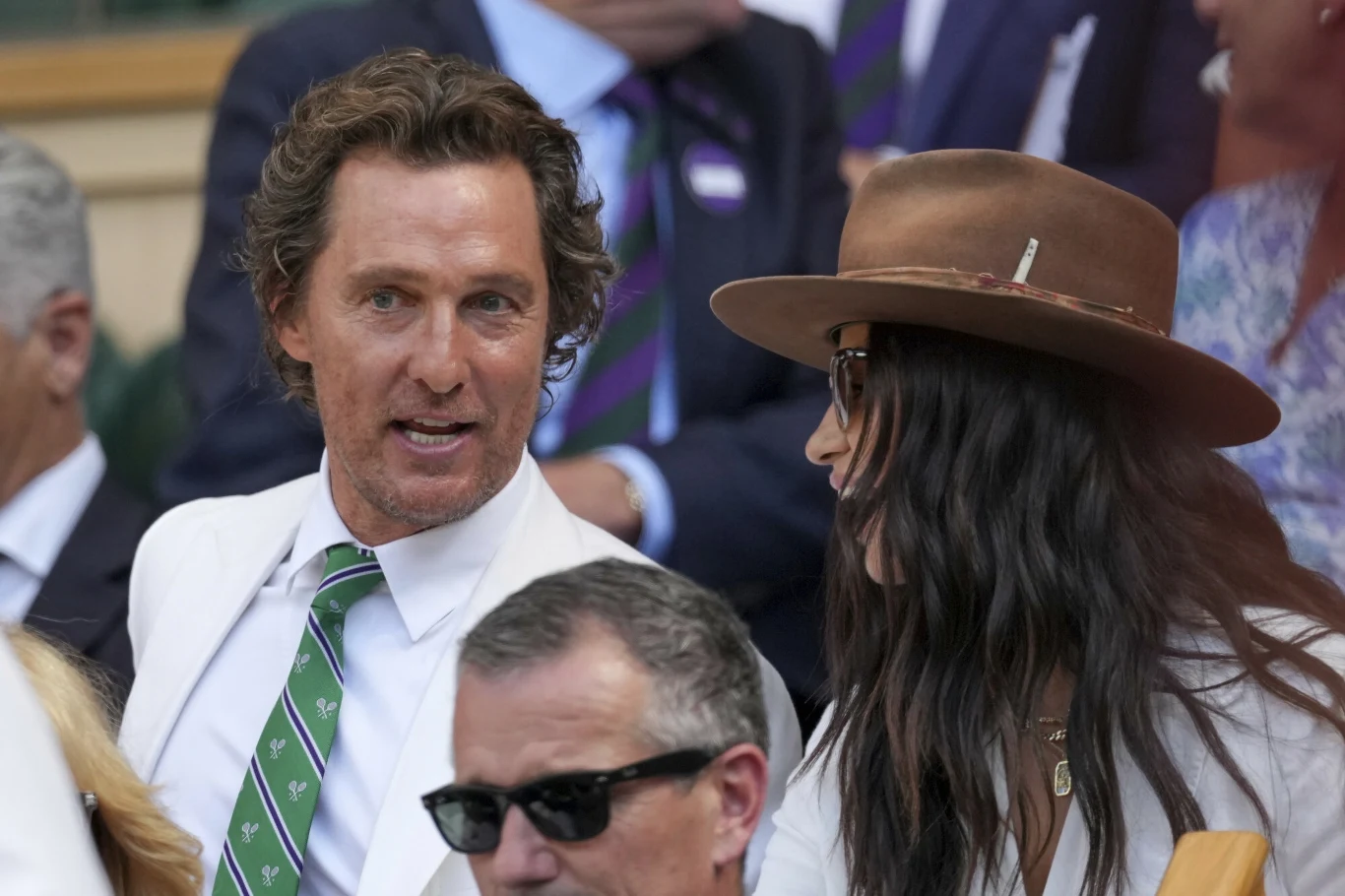 Matthew McConaughey z żoną Camilą Alves na Wimbledonie Mężczyzna w białej marynarce i zielonym krawacie rozmawia z kobietą w kapeluszu, oboje siedzą wśród innych widzów na widowni podczas wydarzenia.