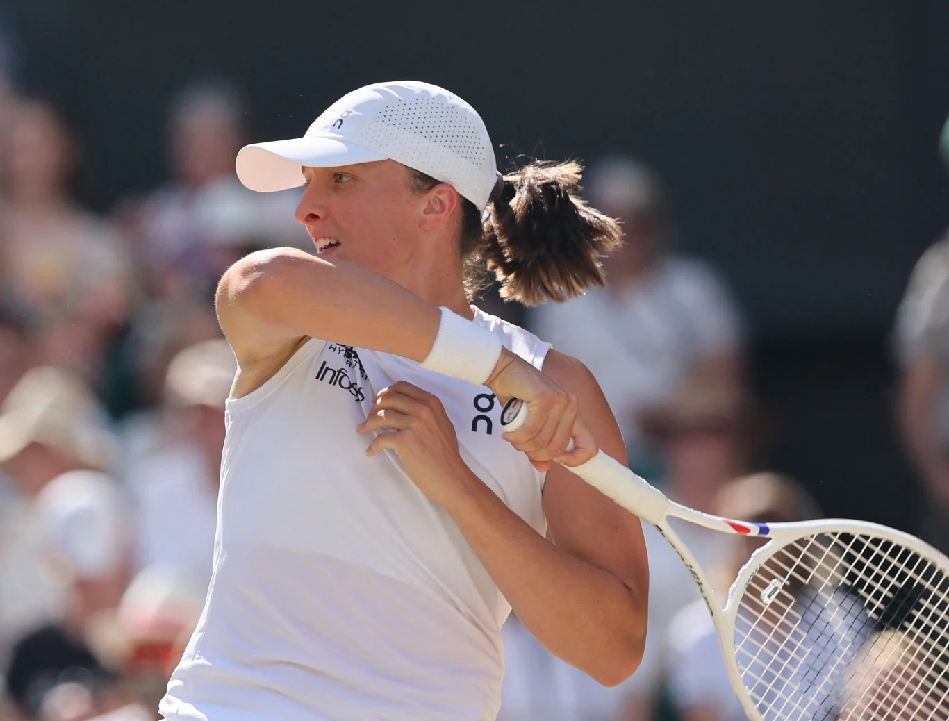 Zbliżenie na tenisistkę w białej sportowej odzieży i czapce z daszkiem, wykonującą dynamiczny ruch rakietą podczas meczu na tle rozmytej publiczności.