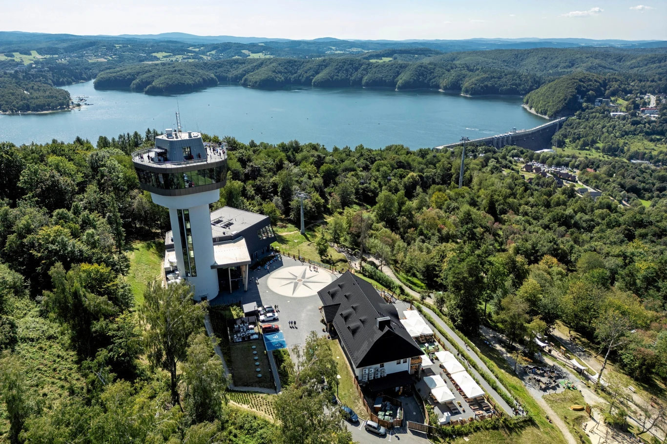 W Solinie otwarto nową atrakcję. Dołącza do bogatej oferty turystycznej miejscowości. Wieża widokowa pośród zielonych drzew, obok niej nowoczesny budynek z czarnym dachem, w tle rozległe jezioro otoczone lasami i wzgórzami, a po prawej stronie widoczna długa zapora wodna.