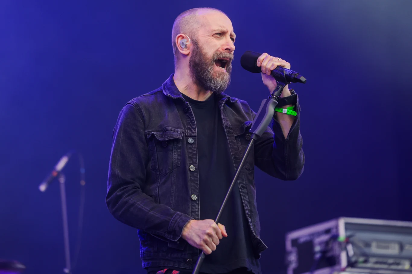 Nick Holmes (Paradise Lost) podczas Mystic Festival 2024 w Gdańsku Brodaty mężczyzna w czarnej kurtce śpiewa do mikrofonu na scenie, w tle widoczny sprzęt muzyczny i oświetlenie sceniczne, atmosfera koncertowa podkreślona kolorowym światłem.