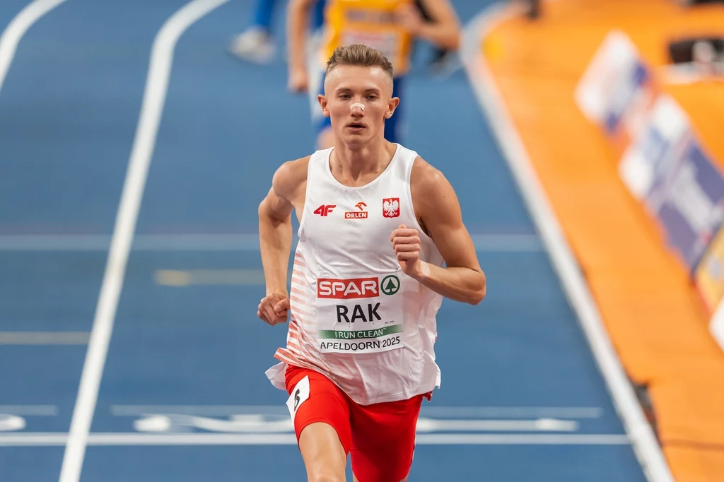 Filip Rak Młody lekkoatleta w biało-czerwonym stroju biegnie po torze na hali sportowej, na piersi ma numer startowy z nazwiskiem i logo zawodów, w tle widoczni inni zawodnicy oraz pomarańczowa nawierzchnia.