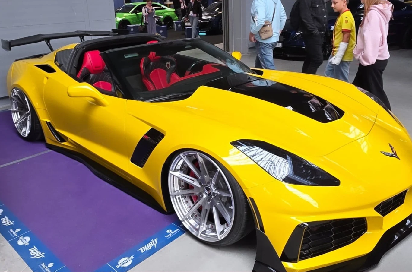 Żółty sportowy samochód typu kabriolet z agresywną stylistyką karoserii, zwracającą uwagę dużymi felgami i niskim zawieszeniem, stoi na targach motoryzacyjnych. Wnętrze pojazdu w dwóch odcieniach czerwieni dodaje mu ekskluzywności, wokół samochodu prze...