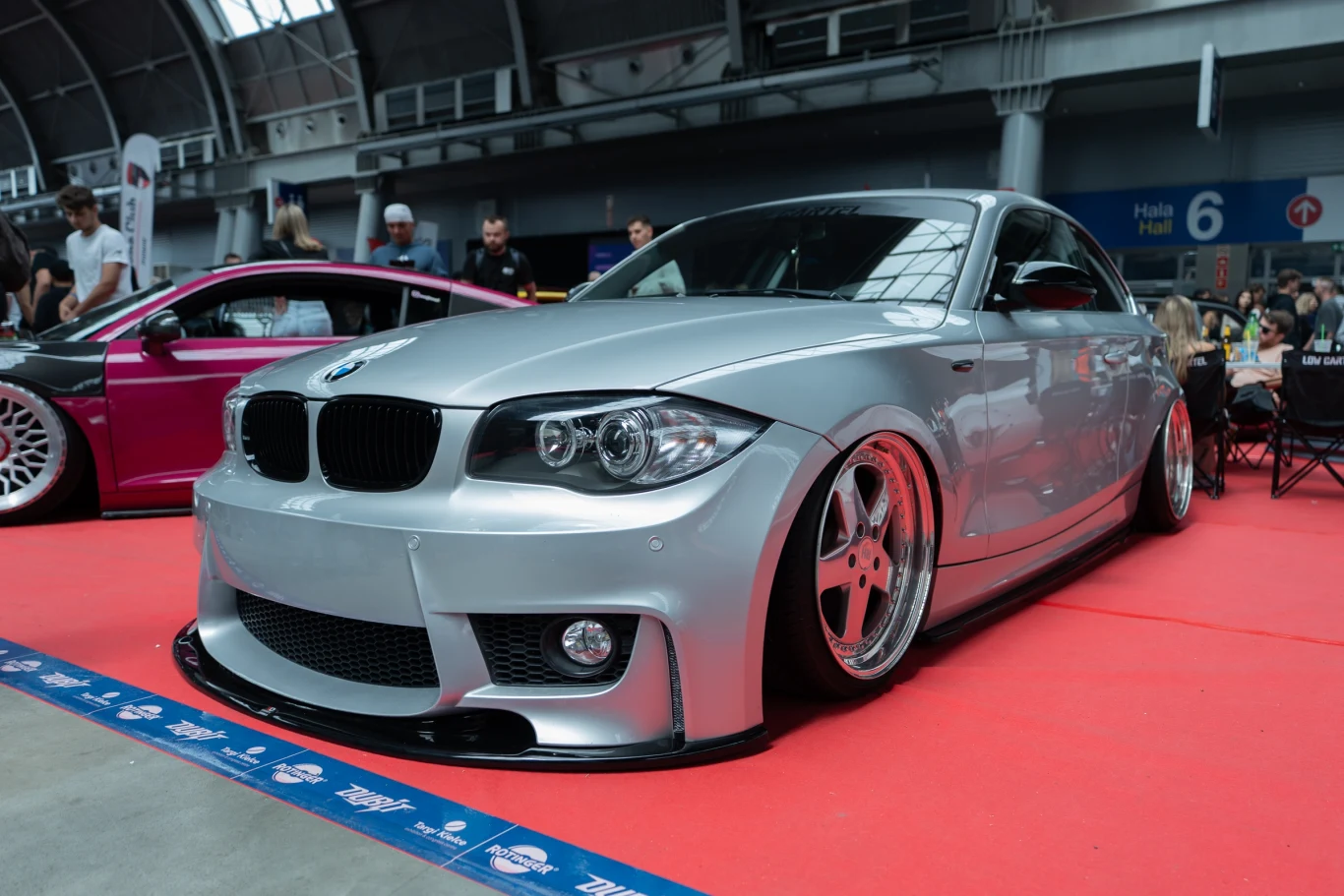 Srebrne sportowe BMW z obniżonym zawieszeniem i szerokimi felgami stoi na czerwonej wykładzinie w hali targowej, w tle widoczni ludzie i inne samochody.