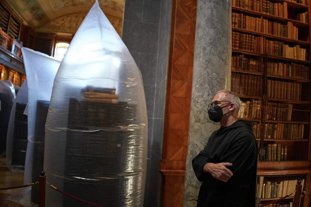 Mężczyzna w maseczce ochronnej stoi w zabytkowej bibliotece, gdzie regały z książkami zabezpieczone są przezroczystymi foliowymi osłonami.