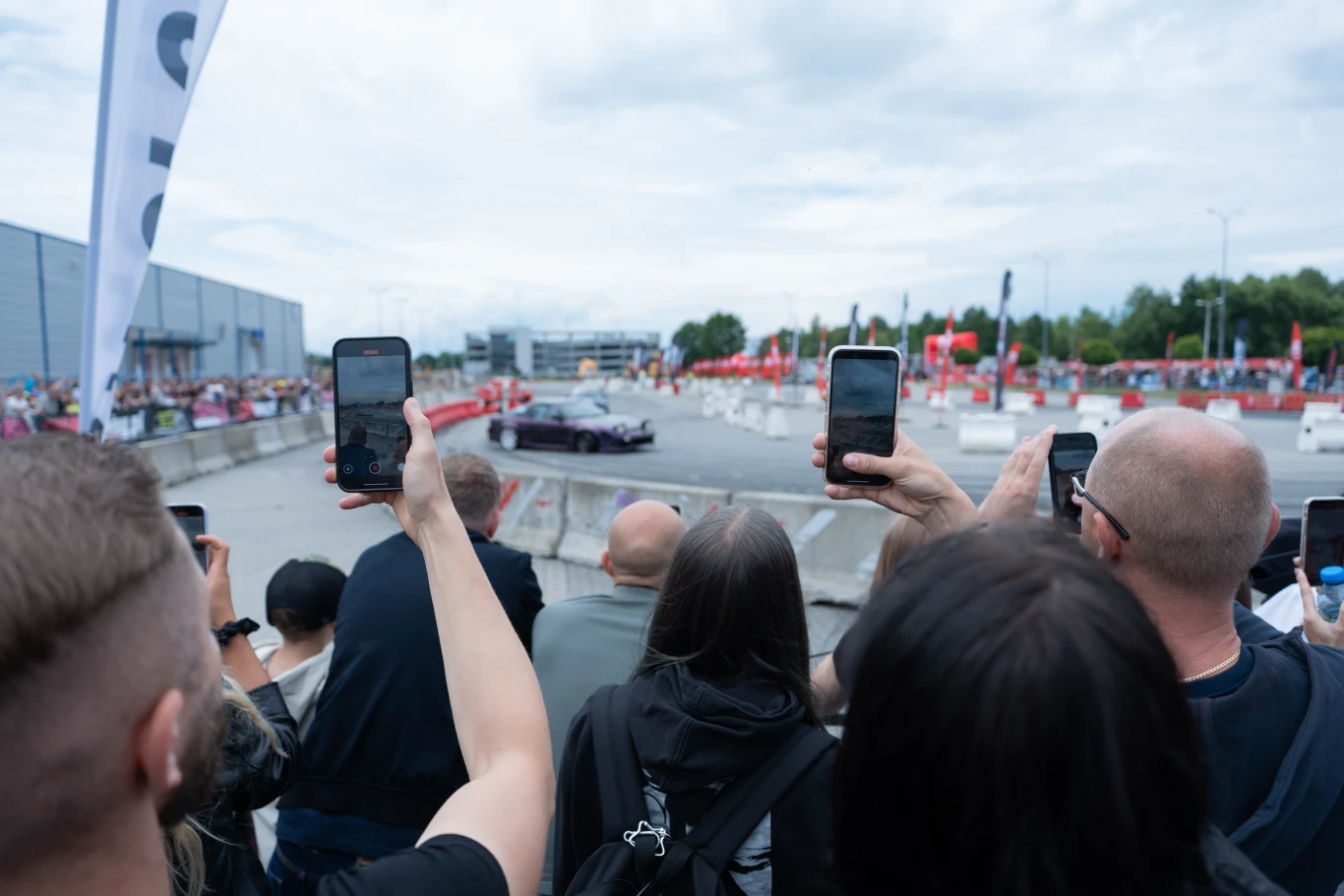 Publiczność skupiona wokół toru wyścigowego z uniesionymi telefonami komórkowymi, nagrywająca przejeżdżający sportowy samochód podczas wydarzenia motoryzacyjnego; w tle widoczne są barierki bezpieczeństwa, flagi oraz zabudowania.