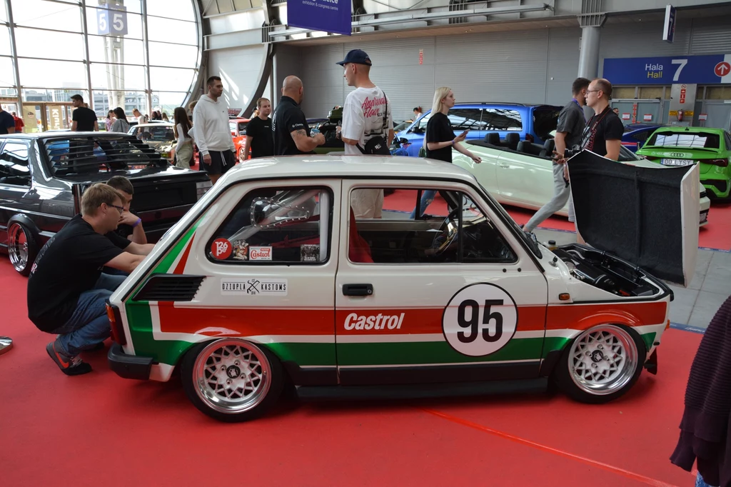 Strefa PRL – powrót do motoryzacyjnych korzeni. Maluch w sportowej wersji z numerem 95 w barwach Castrol, otoczony przez uczestników wydarzenia motoryzacyjnego na hali wystawienniczej, w tle inne samochody tuningowane i osoby oglądające pojazdy.