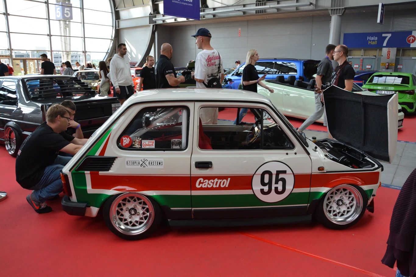 Maluch w sportowej wersji z numerem 95 w barwach Castrol, otoczony przez uczestników wydarzenia motoryzacyjnego na hali wystawienniczej, w tle inne samochody tuningowane i osoby oglądające pojazdy.