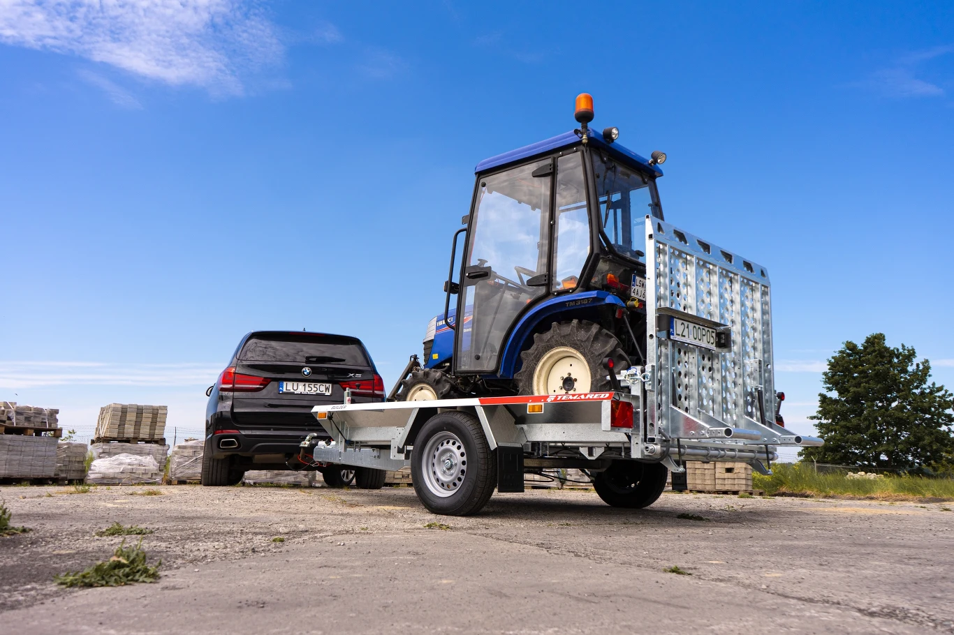 Niebieski traktor na przyczepie podłączonej do czarnego samochodu terenowego, całość ustawiona na betonowej powierzchni, w tle widoczne palety i niskie drzewa, niebo bezchmurne.