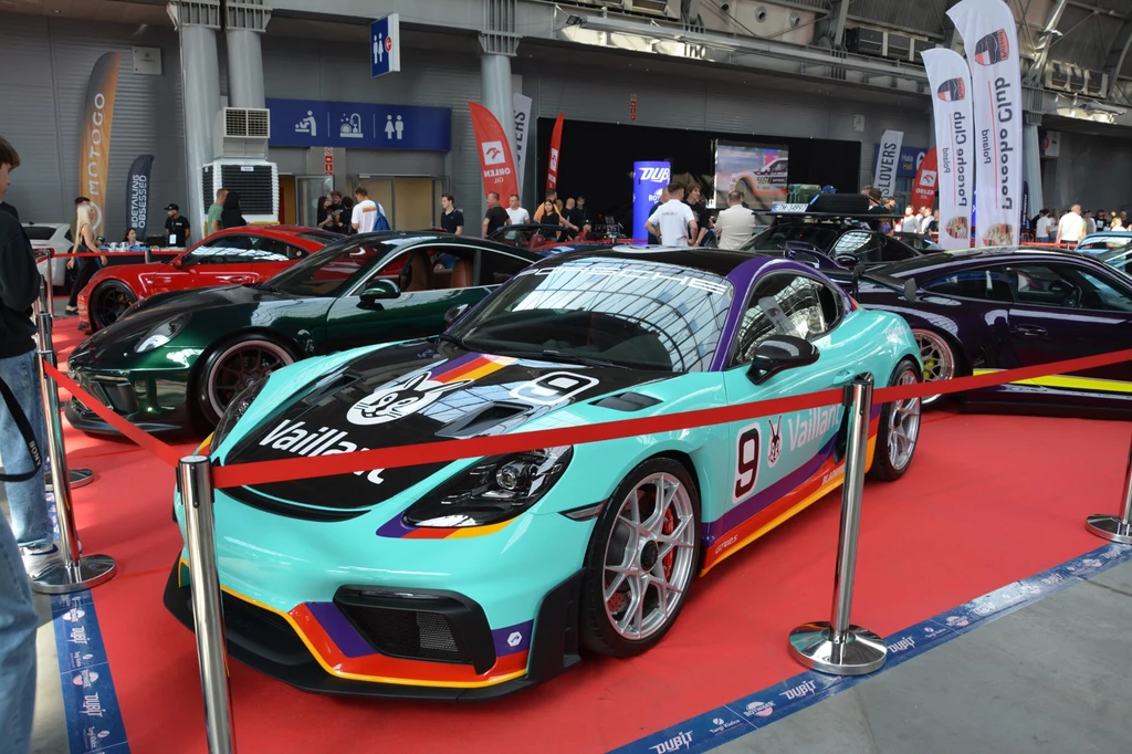 W strefach nie zabrakło też sportowych i szybkich Porsche. Sportowy samochód wyścigowy w kolorach turkusowym i fioletowym z dużym numerem 9 na masce, ustawiony za barierką na czerwonym dywanie podczas targów motoryzacyjnych, w tle kilka innych luksusowych aut oraz odwiedzający imprezę.