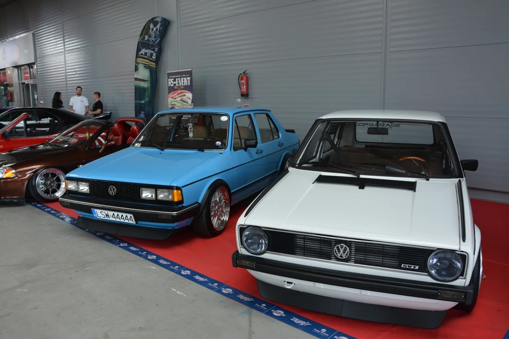 Sekcja dla nostalgików klasycznej motoryzacji. Dwa klasyczne samochody marki Volkswagen stojące obok siebie na wystawie motoryzacyjnej, jedno niebieskie, drugie białe, w tle widoczni uczestnicy wydarzenia oraz inne auta.