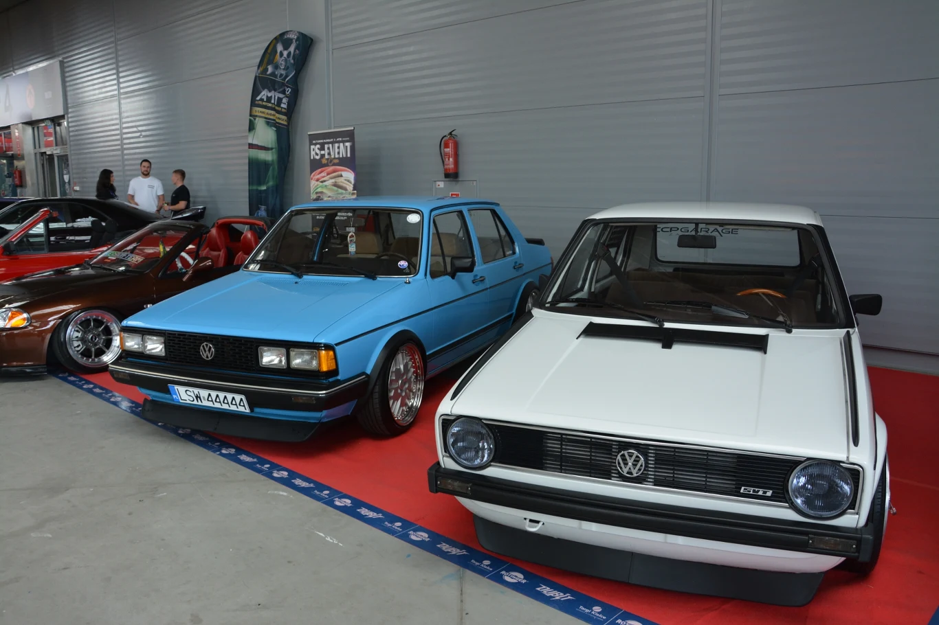 Dwa klasyczne samochody marki Volkswagen stojące obok siebie na wystawie motoryzacyjnej, jedno niebieskie, drugie białe, w tle widoczni uczestnicy wydarzenia oraz inne auta.