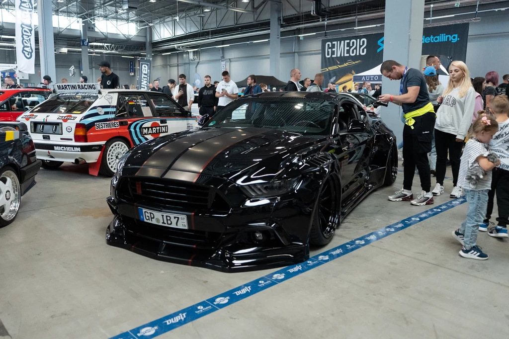 Na zlocie nie mohło zabraknąć nisko osadzonych na ziemii Mustangów. Czarny, sportowy samochód z przyciemnianymi szybami na wystawie motoryzacyjnej, otoczony przez zainteresowanych zwiedzających, w tle klasyczne samochody rajdowe z wyraźnymi napisami sponsorskim i dużą halą wystawową.