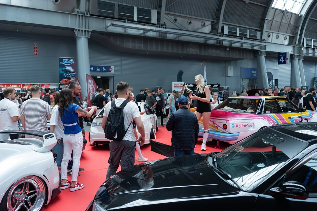Kaido Racer zyskała największą popularność. Tłum ludzi na hali wystawienniczej podziwia sportowe i tuningowane samochody, w centrum uwagi znajduje się kobieta stojąca na podwyższeniu przy kolorowym aucie. Część osób robi zdjęcia, atmosfera jest dynamiczna i skupiona wokół prezentacji motoryzacyj...