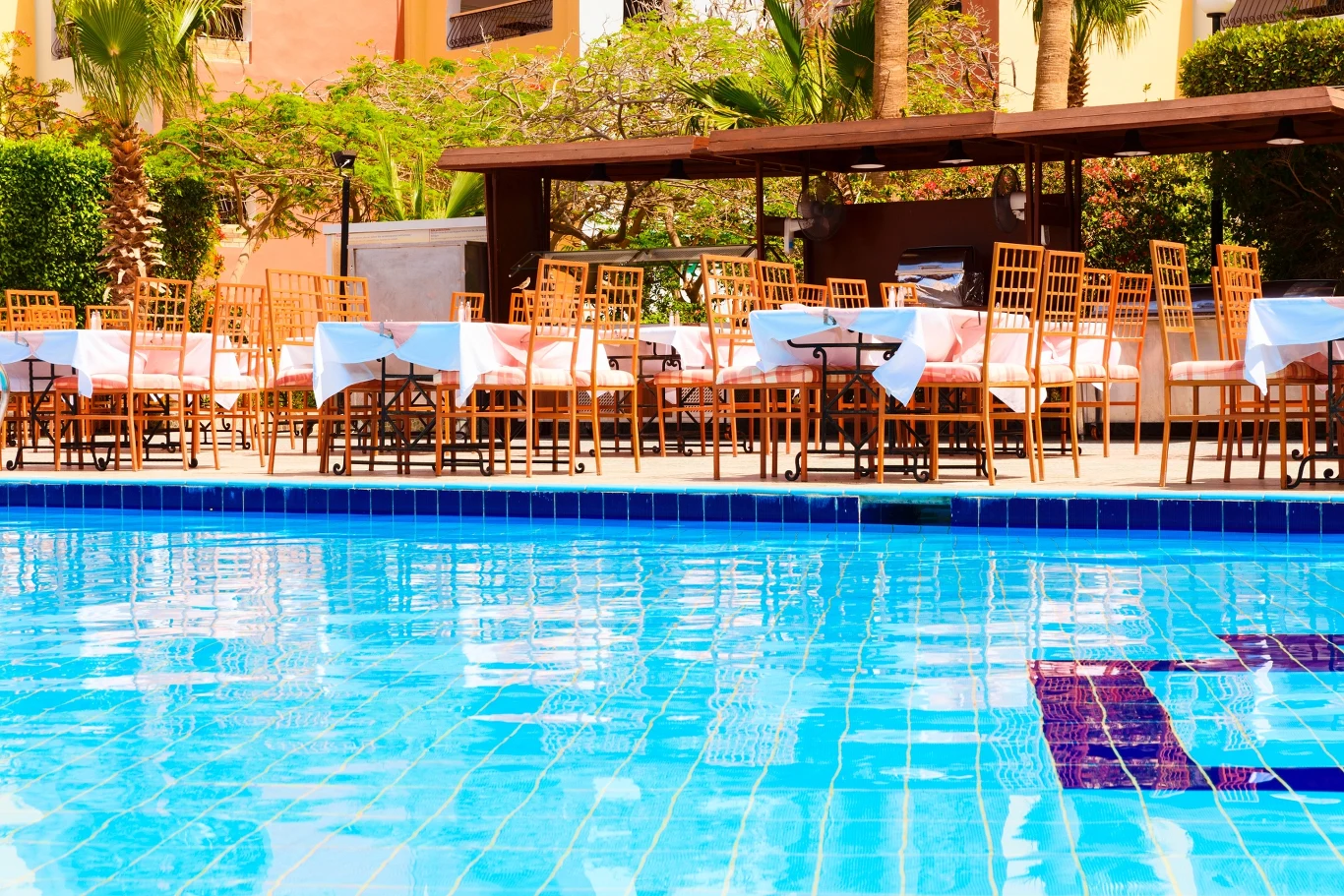 Błękitny basen na pierwszym planie, w tle ustawione elegancko stoliki i krzesła w części restauracyjnej, otoczone palmami i roślinnością, przygotowane na przyjęcie gości.