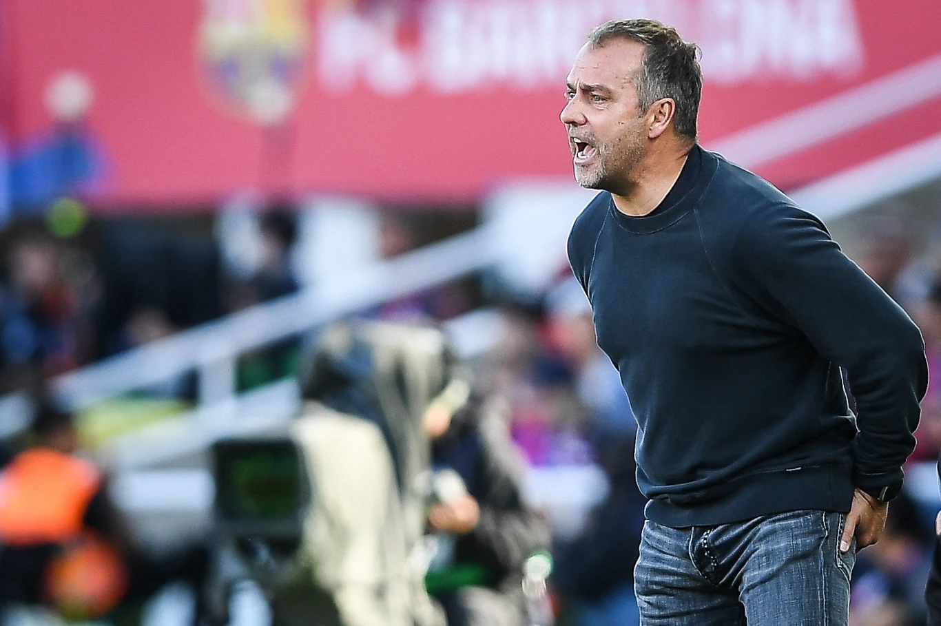 Hansi Flick Mężczyzna w ciemnym swetrze i dżinsach energicznie gestykuluje na tle boiska piłkarskiego, prawdopodobnie w trakcie meczu, w tle rozmazane postacie oraz fragmenty stadionu.