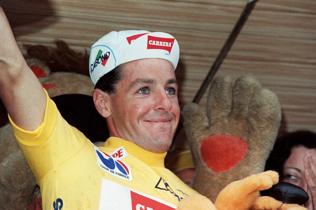 Uśmiechnięty kolarz w żółtej koszulce lidera Tour de France z podniesioną dłonią, w tle maskotka oraz osoby gratulujące zwycięstwa.