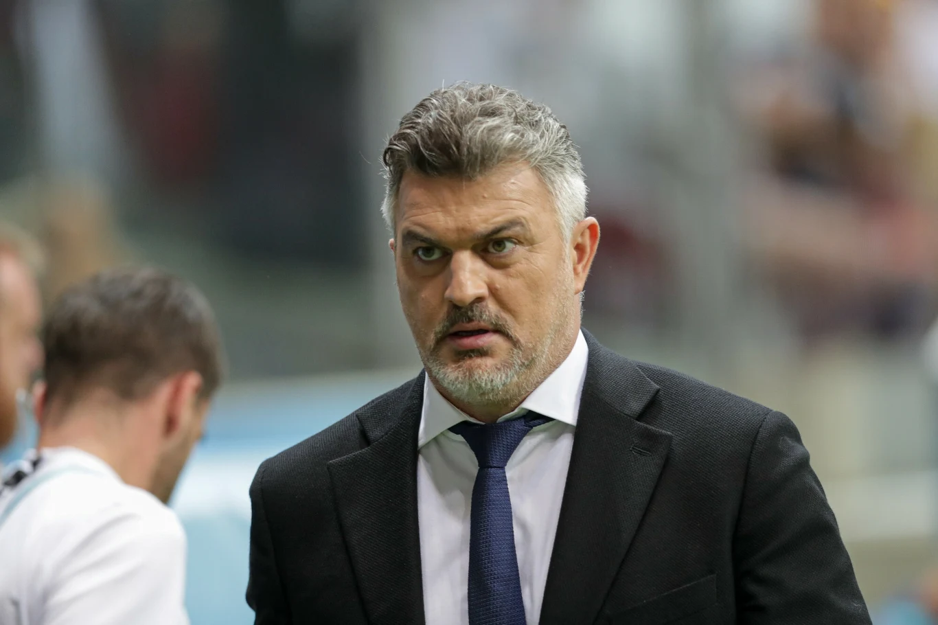 Michał Żewłakow Mężczyzna z krótkimi, siwiejącymi włosami i brodą ubrany w ciemny garnitur, białą koszulę i granatowy krawat, stoi w otoczeniu innych osób w tle, z poważnym wyrazem twarzy.