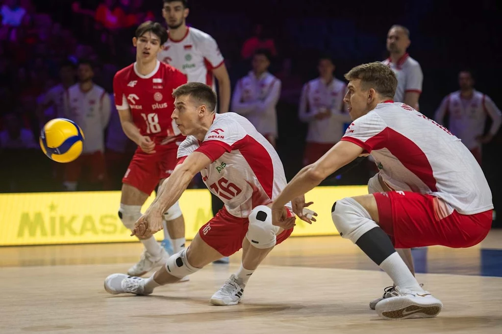 Kamil Semeniuk i Artur Szalpuk Zawodnicy drużyny siatkarskiej ubrani w czerwono-białe stroje wykonują dynamiczną akcję odbioru piłki podczas meczu na hali sportowej, koncentrując się na piłce lecącej nisko nad podłogą.