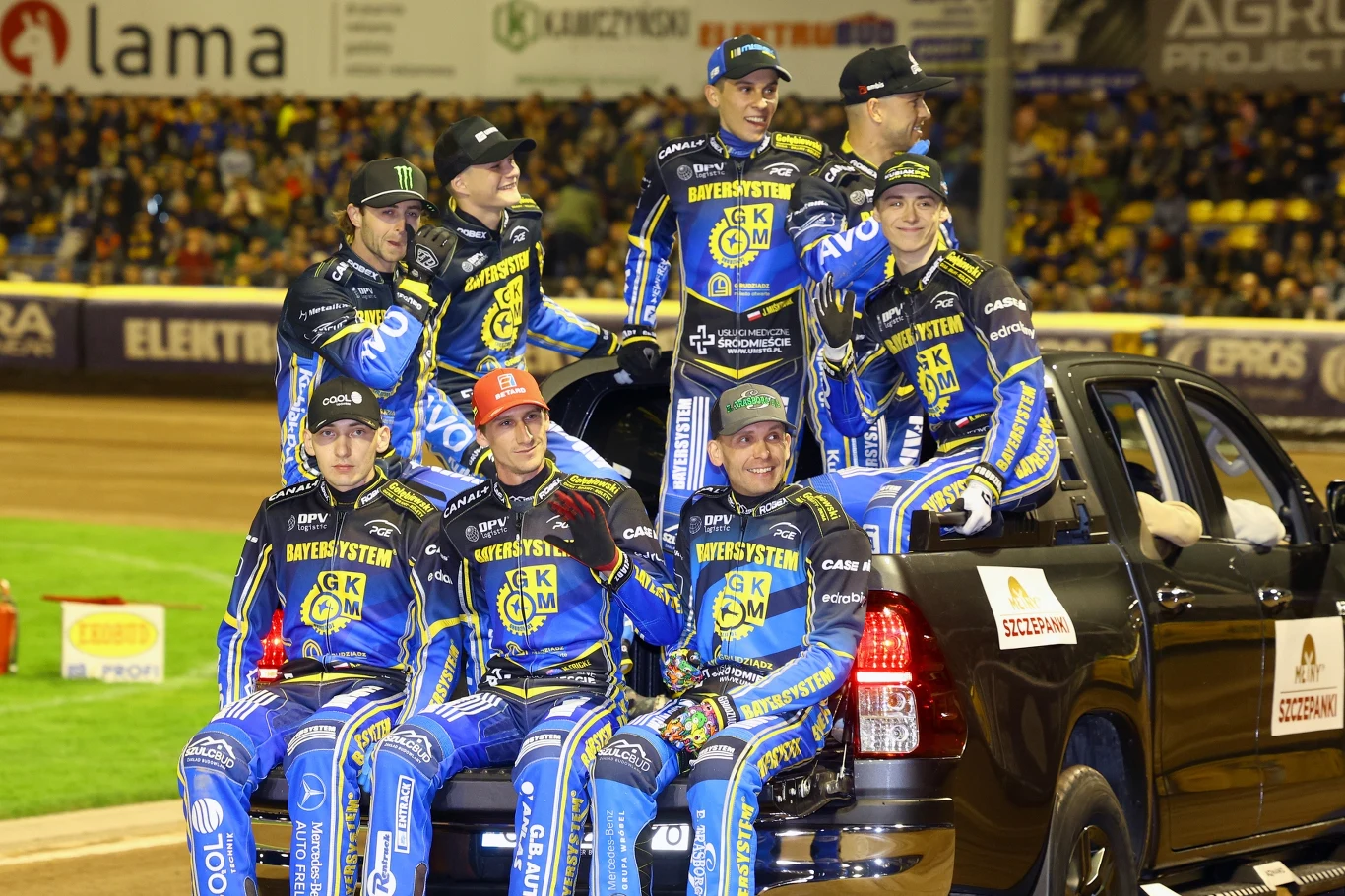 Grupa zawodników żużlowych w niebiesko-żółtych kombinezonach zespołu GKM Grudziądz siedzi i stoi w otwartym bagażniku czarnego pickupa na stadionie podczas wydarzenia sportowego; w tle widoczna jest publiczność oraz elementy toru żużlowego.