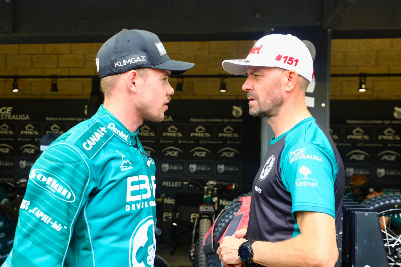 Damian Ratajczak w rozmowie z Piotrem Protasiewiczem Dwóch mężczyzn w sportowych strojach drużynowych prowadzi rozmowę twarzą w twarz w otoczeniu zaplecza technicznego, prawdopodobnie podczas wydarzenia sportowego.