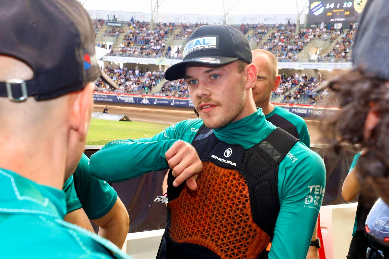 Damian Ratajczak Młody mężczyzna w sportowym stroju i czapce z daszkiem, otoczony przez innych członków drużyny, stoi na stadionie żużlowym podczas zawodów, widząc za plecami trybuny pełne kibiców.