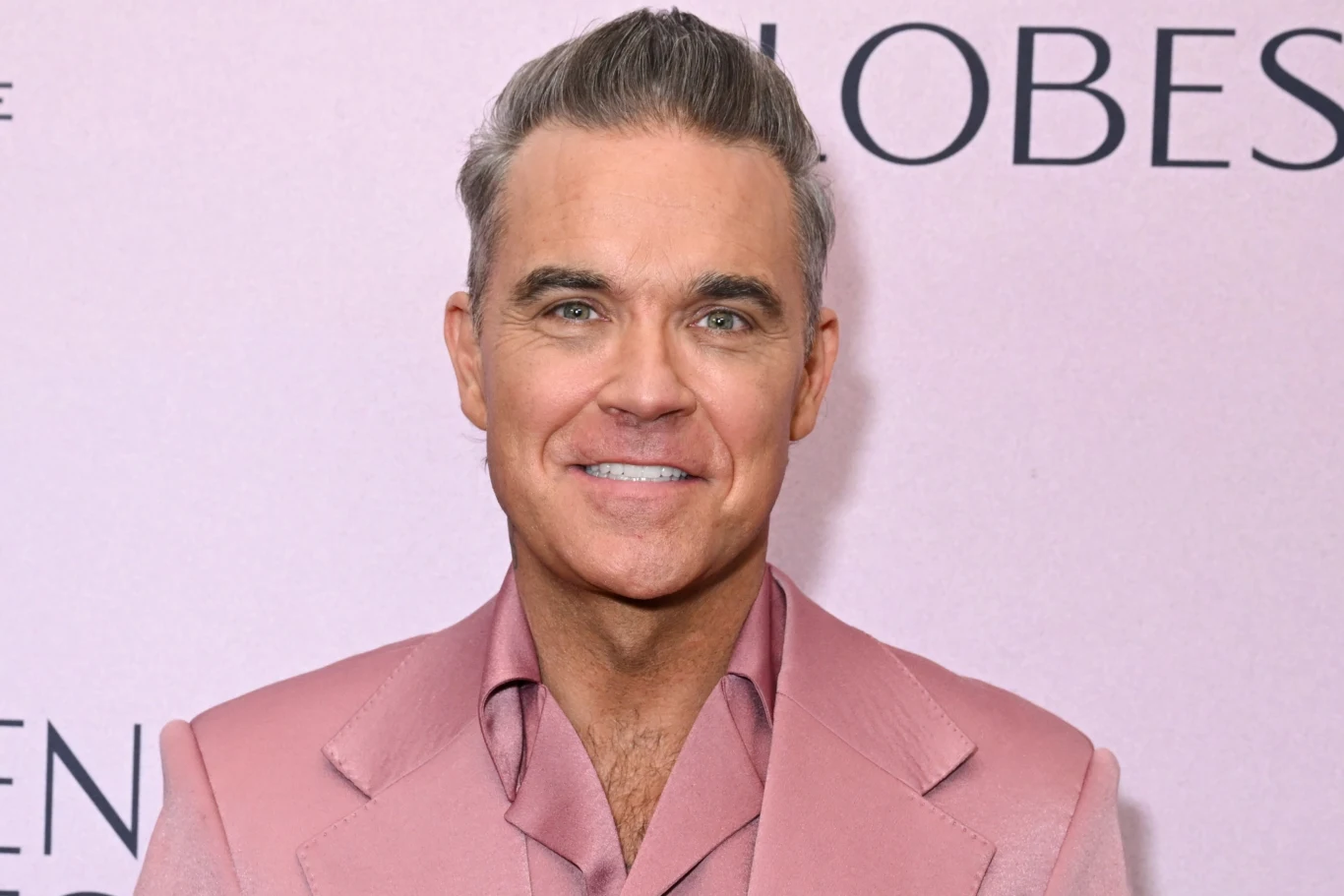 Robbie Williams Mężczyzna w średnim wieku ubrany w różowy garnitur, stojący na tle jasnoróżowej ścianki z częściowo widocznymi napisami, uśmiecha się w kierunku aparatu.