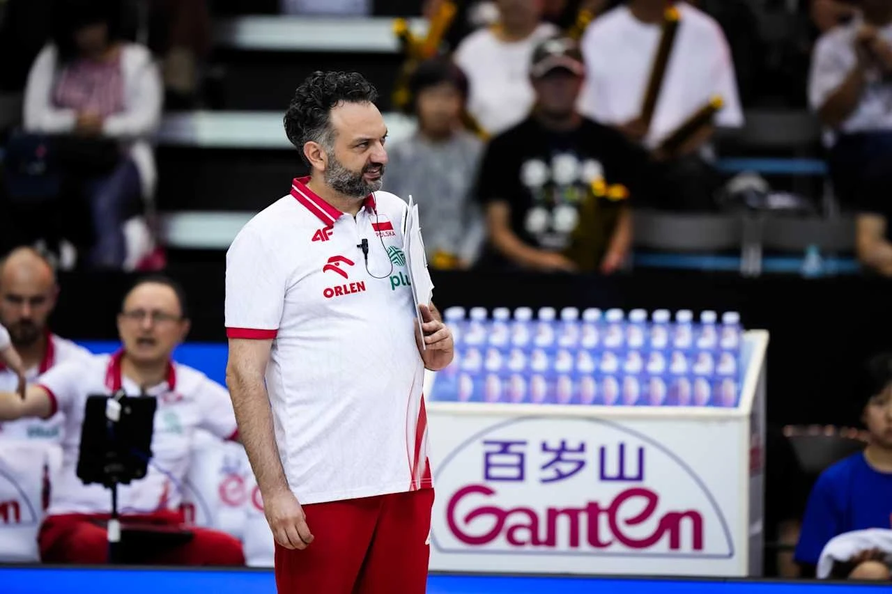 Trener siatkówki w białej koszulce z czerwonymi akcentami oraz logotypem ORLEN i napisem POLSKA stoi na hali sportowej, trzymając notatnik, w tle widoczni są inni członkowie drużyny oraz publiczność.