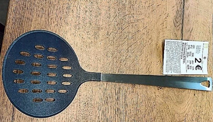 Okrągła cedzakowa łyżka z metalową rączką, umieszczona na drewnianym blacie, obok znajduje się metka z ceną 2 euro.