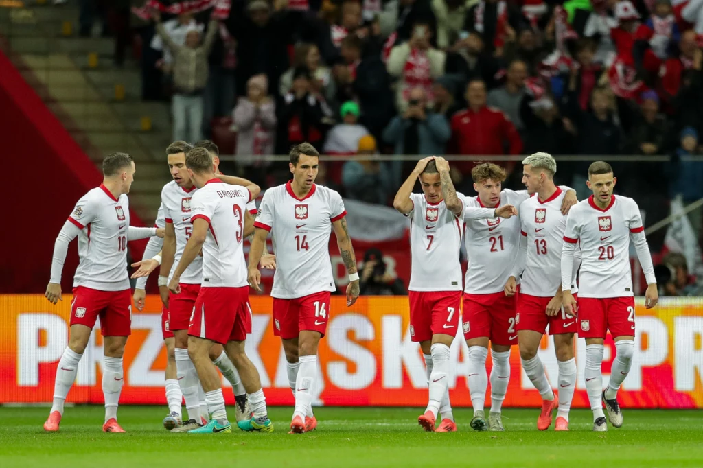 Reprezentacja Polski Grupa polskich piłkarzy w białych koszulkach z czerwonymi elementami i numerami, stojących na murawie stadionu podczas meczu, w tle kibice na trybunach.