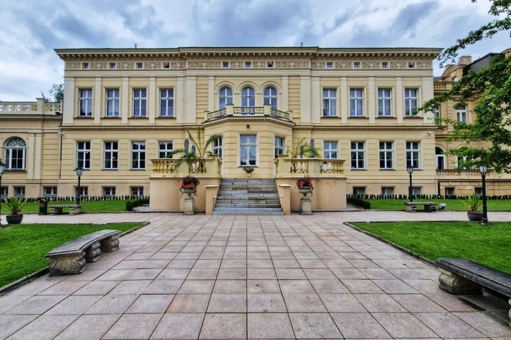 Pałac Nowy widziany od strony parku Zabytkowy pałac z jasną elewacją, licznymi oknami oraz ozdobnymi detalami architektonicznymi, szerokie schody prowadzące do wejścia głównego, po bokach egzotyczne rośliny w dużych donicach, zadbany brukowany dziedziniec otoczony zielonym trawnikiem i k...