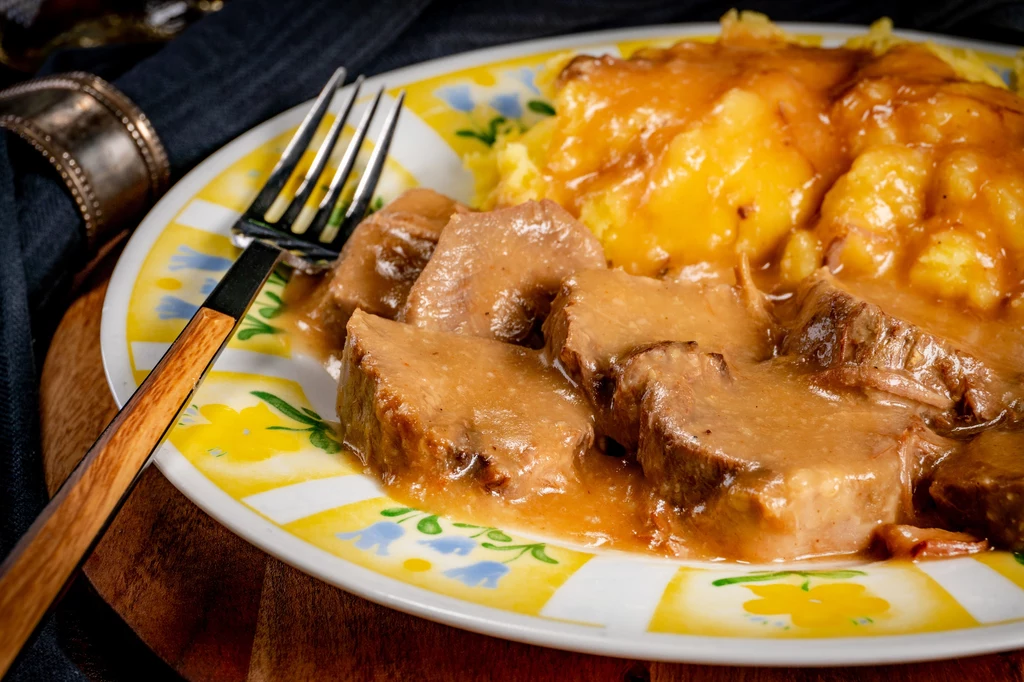 Schab w aromatycznym sosie to uczta dla podniebienia Kawałki wołowiny polane gęstym brązowym sosem obok porcji ziemniaczanego purée na kolorowym talerzu, widoczny widelec po lewej stronie talerza.