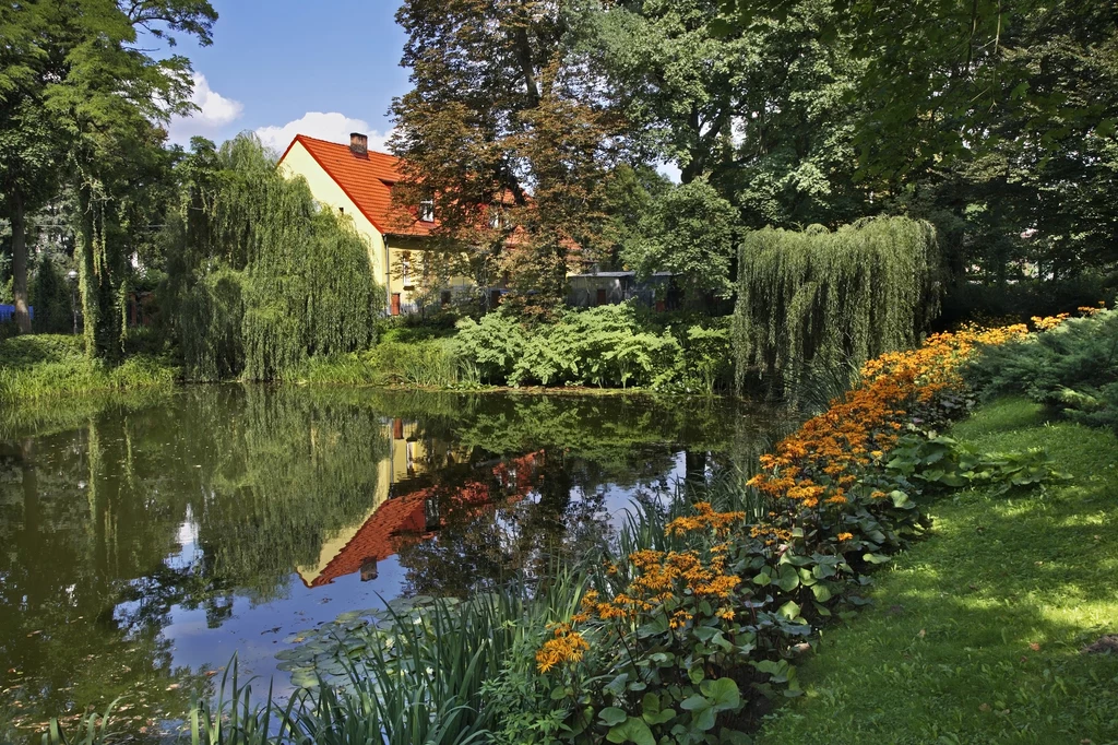 Park otaczający pałace w Ostromecku to miejsce idealne na spacer lub piknik Dom o czerwonym dachu otoczony bujną zielenią i drzewami, przed którym znajduje się staw z odbiciem budynku oraz kolorowe rabaty kwiatowe i przycięta trawa.