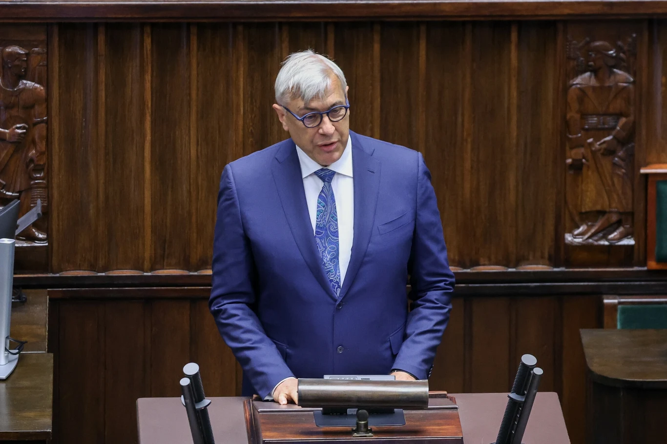 Roman Giertych zwrócił się w liście do biskupów. Zdjęcie ilustracyjne Mężczyzna w garniturze i okularach stoi za mównicą w sali obrad, w tle drewniany wystrój oraz rzeźbione elementy ściany.