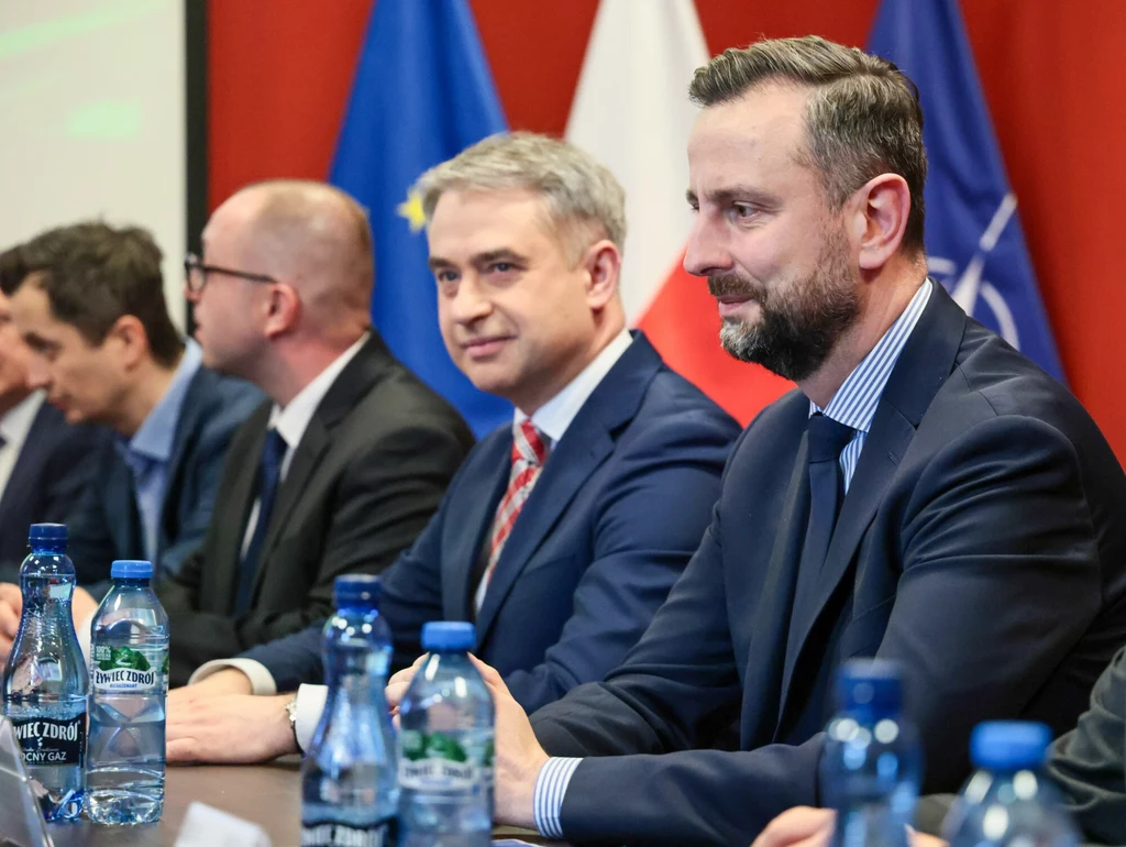 Wicepremierzy Krzysztof Gawkowski i Władysław Kosiniak-Kamysz Grupa mężczyzn w garniturach siedzących przy stole konferencyjnym, na stole stoją butelki z wodą, w tle widoczne flagi Polski, Unii Europejskiej i NATO.