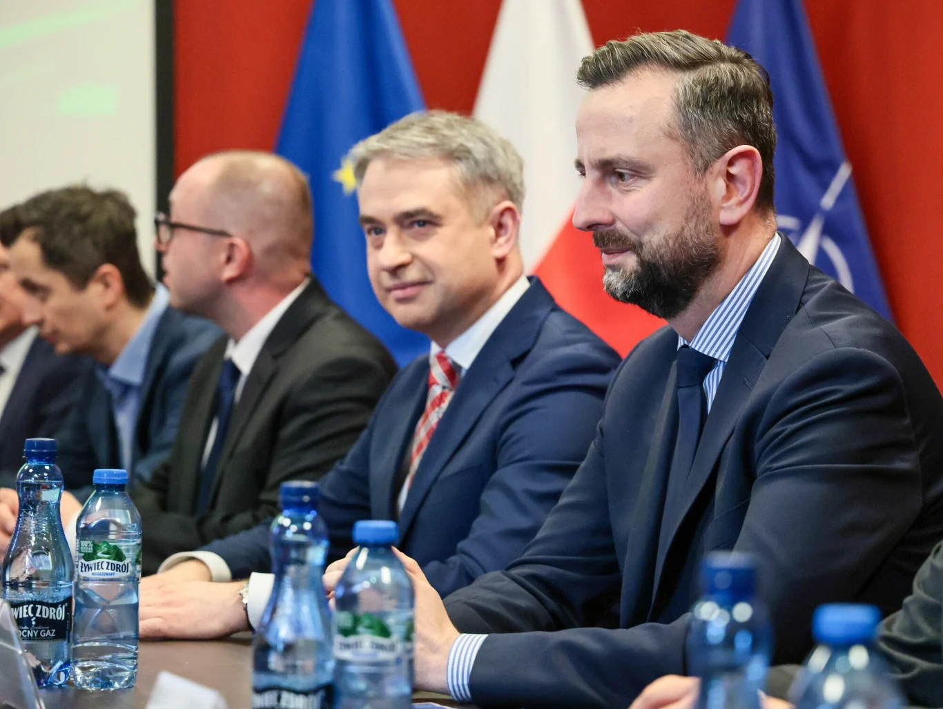 Grupa mężczyzn w garniturach siedzących przy stole konferencyjnym, na stole stoją butelki z wodą, w tle widoczne flagi Polski, Unii Europejskiej i NATO.