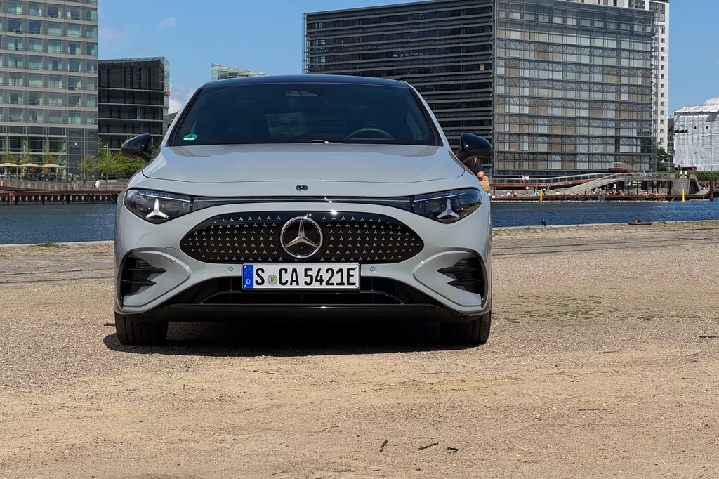 Przód auta przeładowano "gwiazdami", znajdziemy je nawet w światłach do jazdy dziennej Nowoczesny, srebrny samochód marki Mercedes zaparkowany na tle miejskiej architektury z przeszklonymi biurowcami oraz wodą w oddali.
