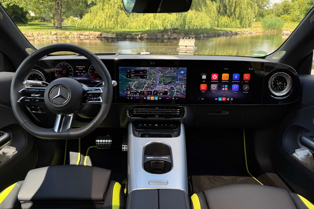 W kokpicie dominuje wielki panel skrywający 3 ekrany Wnętrze nowoczesnego samochodu marki Mercedes z widocznym logo na kierownicy, cyfrowymi wyświetlaczami na desce rozdzielczej, systemem multimedialnym z mapą na ekranie centralnym oraz skórzanymi fotelami z żółtymi akcentami, w tle przez szybę widoczny ...