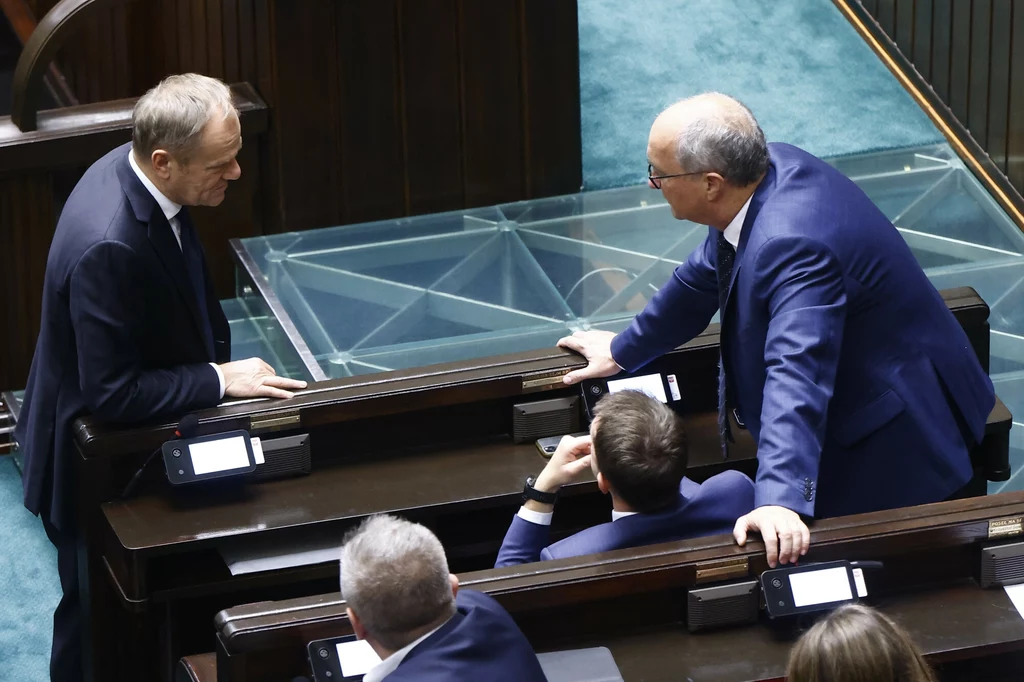 Premier Donald Tusk i wicemarszałek Sejmu Włodzimierz Czarzasty Trzech elegancko ubranych mężczyzn w garniturach prowadzi ożywioną rozmowę przy ławach sejmowych, w tle widać fragment nowoczesnego wnętrza parlamentu.